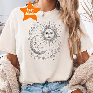 May include: A cream-colored short-sleeve t-shirt featuring a black line-art graphic of a sun and crescent moon with human faces, surrounded by stars and leafy vines. The shirt has a crew neckline and is worn with blue denim shorts.