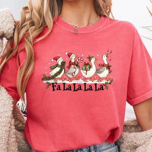 May include: A coral-colored short-sleeve t-shirt featuring a graphic of four geese wearing winter accessories like scarves and hats. The geese are carrying holiday items including a wreath, candy cane, and gift. The text Fa La La La La is printed below.