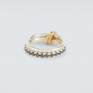 May include: A multi-band ring featuring a polished silver-tone band set with a row of small, round, black stones and a separate, thinner, smooth gold-tone band that loops through the silver band, creating a layered and interconnected design.