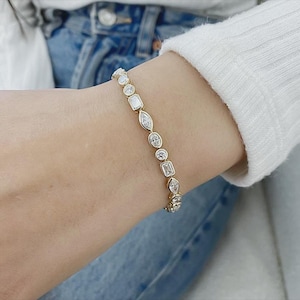 May include: A gold-toned bracelet featuring a series of bezel-set clear stones in alternating shapes, including round, marquise, and emerald cuts. The stones are linked together in a continuous line, creating a refined and structured design.