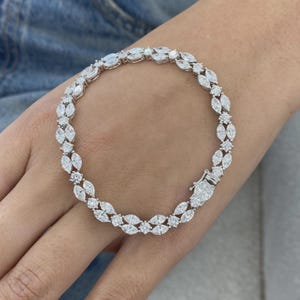 Op de afbeelding: Een zilverkleurige tennisarmband met een herhalend patroon van heldere stenen in marquise- en ronde slijpvorm. De armband is voorzien van een metalen bakslot en heeft een flexibele schakelstructuur voor een soepele pasvorm.