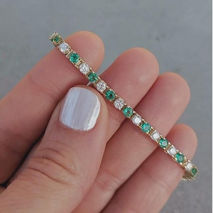 Op de afbeelding: Een gouden tennisarmband met een afwisselend patroon van rondgeslepen groene smaragden en heldere witte diamanten in een doorlopende lijn. De dunne, gepolijste metalen band heeft een klassiek ontwerp met gelijkmatig verdeelde edelstenen.