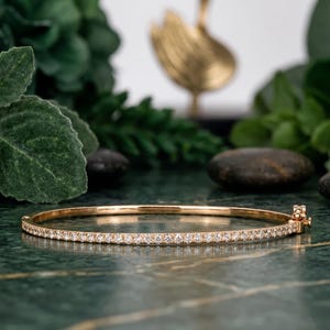 Może przedstawiać: Cienka złota bransoletka typu bangle z ciągłym rzędem małych, przezroczystych, okrągłych kamieni osadzonych wzdłuż przedniej połowy obręczy. Bransoletka posiada mały, bezpieczny mechanizm zapięcia na zawiasie umieszczony z boku.