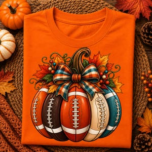 May include: An orange crew neck t-shirt featuring a graphic of a pumpkin constructed from several American footballs in shades of brown, orange, cream, and teal. The pumpkin is adorned with a plaid bow, autumn leaves, and small berries at the top.