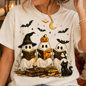 Peut inclure: Un t-shirt couleur crème avec un motif représentant trois fantômes blancs lisant des livres. Un fantôme porte un chapeau de sorcière, un autre une citrouille sur la tête, et le troisième une chauve-souris. Un chat noir est assis à côté.