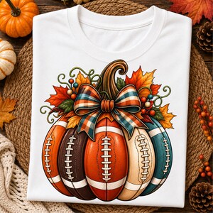 May include: A white t-shirt featuring a graphic of a pumpkin constructed from segments resembling American footballs in orange, brown, and teal colours. The pumpkin is adorned with a plaid bow, autumn leaves, and berries, creating a seasonal design.
