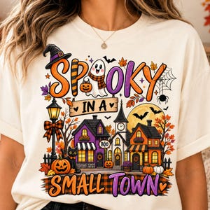 Op de afbeelding: Een crèmekleurig t-shirt met een kleurrijke afbeelding van een dorp in Halloween-thema met de tekst "SPOOKY IN A SMALL TOWN". Het ontwerp bevat een geest, een zwarte kat, pompoenen, een lantaarnpaal, huizen, vleermuizen en herfstbladeren.