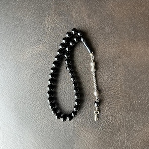 Peut inclure: Un chapelet composé de perles noires, lisses et rondes, disposées en boucle. Le cordon est relié à un élément décoratif en métal argenté comprenant une pièce cylindrique texturée, une petite perle noire et un pendentif en métal argenté.