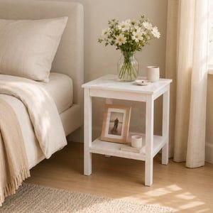 May include: A white wooden side table with a square top and a lower shelf. The top holds a clear glass vase filled with white daisies and a small beige cup. The lower shelf displays a framed photograph and a small white cylindrical container.