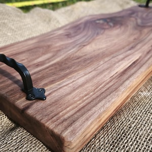 May include: A rectangular wooden serving tray featuring a natural live edge and a rich brown wood grain pattern. The tray is equipped with two black metal handles, each with a twisted design, secured by screws at both ends of the board.