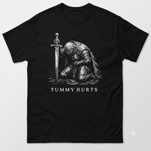 May include: A black t-shirt featuring a white graphic illustration of a knight in full armour kneeling and holding a sword. Below the knight, the text "TUMMY HURTS" is printed in a clean, white, sans-serif font centred on the chest area.