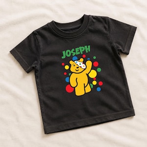 May include: A black short-sleeved t-shirt featuring a graphic of a yellow bear with a polka-dot eye patch, surrounded by colourful circles. The name JOSEPH is printed in green capital letters above the bear design on the front of the shirt.