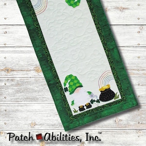 May include: A green and white table runner featuring a gnome and pot of gold design. The table runner measures 16 inches by 37 1/2 inches and is labelled "Lucky Gnome Y'are" and "St. Pat's Table Runner".
