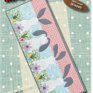 Könnte beinhalten: Ein Quiltmuster für einen Osterhasen-Läufer. Das Muster zeigt eine Reihe von weißen Hasen mit grauen Ohren und floralen Akzenten. Das Muster ist 12 Zoll mal 32 Zoll groß und wird als Anfänger-Level eingestuft.