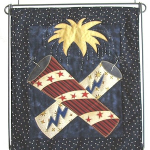 May include: A black and white quilted wall hanging featuring a red, white and blue firework design. The design depicts two crossed fireworks with a yellow starburst above them.