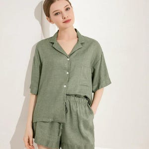 May include: A sage green two-piece pajama set consisting of a short-sleeved button-down shirt with a notched collar and a single chest pocket, paired with matching elastic-waist shorts featuring a drawstring and a wide hem detail.
