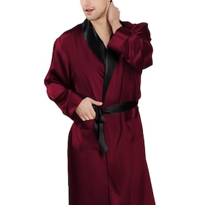 May include: A burgundy robe with a smooth, satin-like finish, featuring a contrasting black shawl collar, long sleeves, and a matching black tie belt at the waist. The garment has a knee-length hem and a clean, simple design with no patterns.