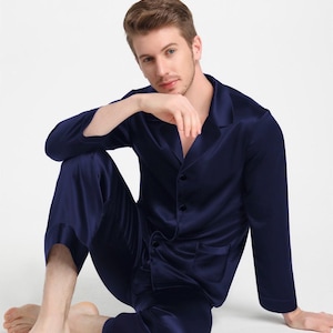 May include: A navy blue pajama set consisting of a long-sleeved button-down shirt with a notched collar and a chest pocket, paired with matching long trousers. The fabric has a smooth, satin-like finish with a subtle sheen throughout the garment.