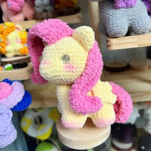 May include: A handmade crochet plush toy shaped like a pony with a pale yellow body and bright pink yarn mane and tail. The figure features two glossy, round, light-colored eyes and soft, textured yarn construction, standing on a small wooden base.