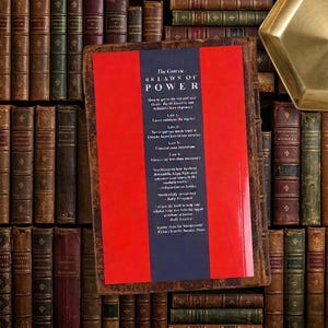 Pu&ograve; includere: Un libro intitolato The Concise 48 Laws of Power con una copertina rossa e blu scuro con testo bianco, posizionato su uno sfondo di numerosi libri antichi rilegati in pelle con dorsi dorati e l'angolo di un vassoio in metallo dorato.