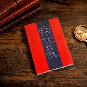 Pu&ograve; includere: Un libro intitolato The Concise 48 Laws of Power con una copertina rosso brillante e una striscia verticale blu scuro al centro contenente testo bianco. Il libro &egrave; posizionato su una superficie in legno scuro accanto a una bussola dorata e libri in pelle.