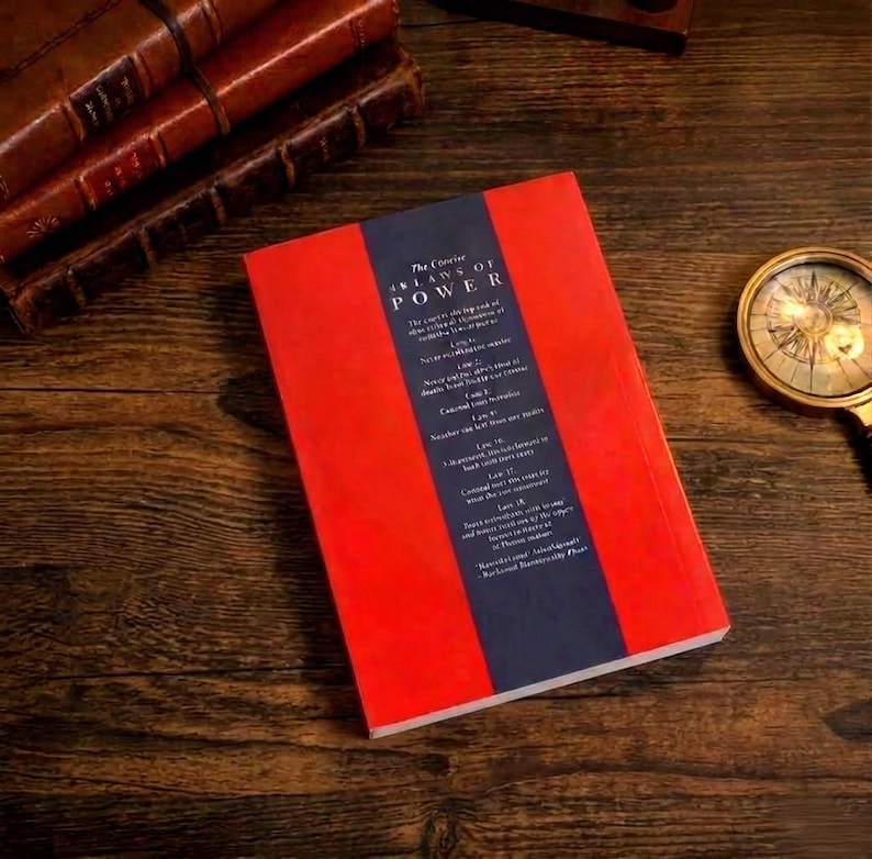 Pu&ograve; includere: Un libro intitolato The Concise 48 Laws of Power con una copertina rosso brillante e una striscia verticale blu scuro al centro contenente testo bianco. Il libro &egrave; posizionato su una superficie in legno scuro accanto a una bussola dorata e libri in pelle.