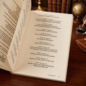 Pu&ograve; includere: Un libro aperto con pagine color crema che mostra un sommario. Il testo elenca le leggi dalla 20 alla 30, inclusi titoli come Do Not Commit to Anyone, Play a Sucker to Catch a Sucker e Make Your Accomplishments Seem Effortless.