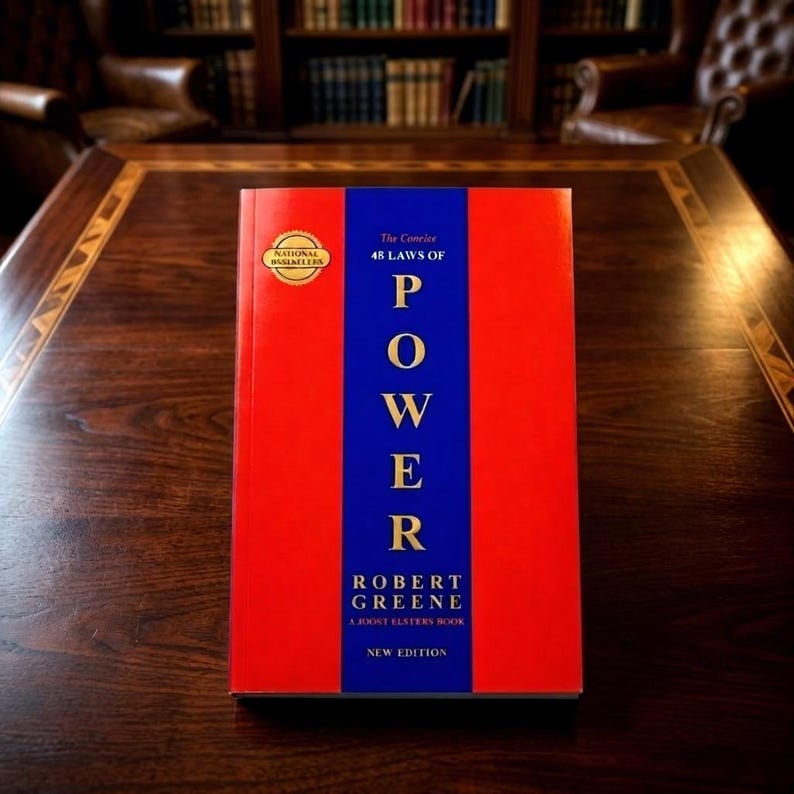 Pu&ograve; includere: Un libro con copertina rigida intitolato The Concise 48 Laws of Power di Robert Greene. La copertina presenta una striscia blu verticale centrata su uno sfondo rosso con testo dorato. Un sigillo dorato in alto a sinistra recita National Bestseller. In basso &egrave; stampato New Edition.