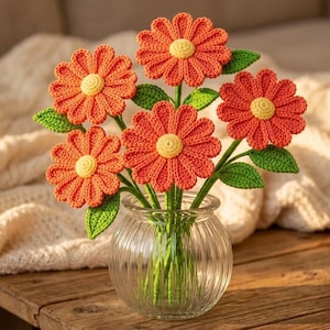 Puede incluir: Un ramo de cinco flores de ganchillo de color naranja con centros amarillos y hojas verdes, dispuesto en un jarrón de cristal transparente y acanalado. Cada flor presenta pétalos texturizados y un disco central, con tallos verdes visibles.