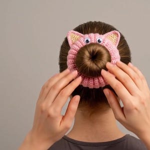 Könnte beinhalten: Ein rosa gehäkeltes Haargummi in Schweinchenform mit zwei kleinen Ohren mit hellgelber Mitte und zwei aufgeklebten Wackelaugen aus Kunststoff. Das kreisförmige Accessoire umschließt einen braunen Haarknoten und weist ein strukturiertes Strickmuster auf.
