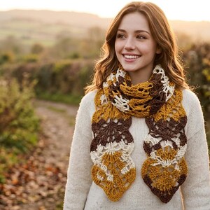 May include: A crochet scarf featuring a repeating leaf pattern in shades of mustard yellow, dark brown, and cream. The scarf is worn over a plain, long-sleeved, off-white knit sweater. The intricate openwork design highlights the yarn texture.