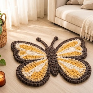 May include: A butterfly-shaped rug featuring a textured, tufted design in shades of dark brown, mustard yellow, and cream. The rug has a central body section with two large upper wings and two smaller lower wings, all outlined in dark brown yarn.