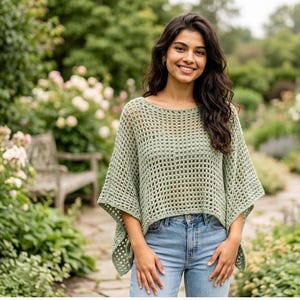 May include: A sage green crochet poncho featuring a wide, open-knit grid pattern, a rounded neckline, and short, loose sleeves, paired with light blue denim jeans. The garment has a boxy, relaxed fit that drapes over the torso and shoulders.