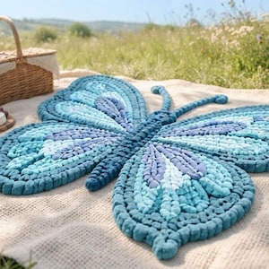 May include: A large, textured rug shaped like a butterfly, crafted from thick yarn in various shades of blue, including navy, sky blue, and teal. The rug features a symmetrical wing pattern with raised, knotted details and two long antennae.