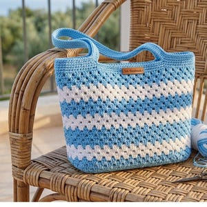 Pode incluir: Uma bolsa de crochê com listras horizontais de fio azul claro e branco em um padrão clássico de quadrados de vovó. A bolsa tem duas alças azuis combinando e uma pequena etiqueta retangular de couro marrom com texto na parte frontal.