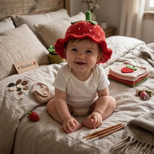 May include: A red crocheted bucket hat with white seed details and a green stem on top. The hat features a wavy brim and is worn by a baby in a white bodysuit. The scene includes small crocheted items, wooden buttons, and three crochet hooks.