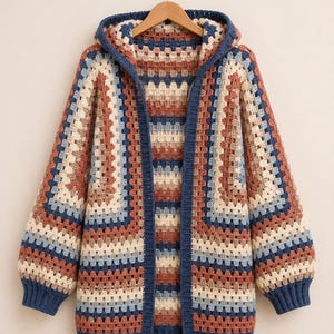 Op de afbeelding: Een vest met lange mouwen en capuchon, gemaakt van gehaakte granny squares in een meerkleurig patroon van roestoranje, crème, lichtblauw en donkerblauw. Het kledingstuk heeft een effen donkerblauwe geribbelde boord langs de opening, mouwen en zoom.