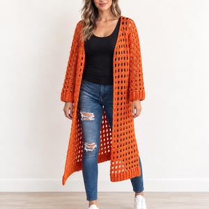 Può includere: Un lungo cardigan arancione all'uncinetto aperto sul davanti, caratterizzato da un motivo geometrico traforato ripetitivo, maniche lunghe con polsini a coste e orlo dritto, indossato sopra una canotta nera, jeans blu strappati e scarpe da ginnastica bianche.