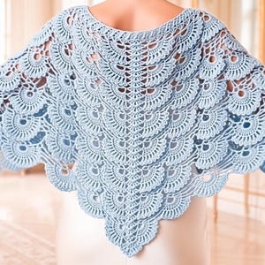 May include: A light blue crochet shawl featuring a repeating fan-shaped pattern. The garment has a wide, triangular silhouette with intricate, openwork stitching that creates a layered, scalloped edge throughout the entire piece.