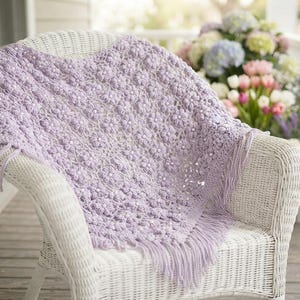 May include: A light lavender crochet blanket featuring a repeating floral pattern with small white beads at the center of each flower. The rectangular textile has a textured surface and is finished with long, matching lavender fringe along the edges.