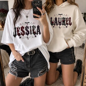 May include: A white t-shirt with the name JESSICA printed in a gothic font featuring red roses, spiderwebs, and a bat, paired with a cream-coloured hoodie with the name LAUREL in a matching gothic style, both worn with black denim shorts.