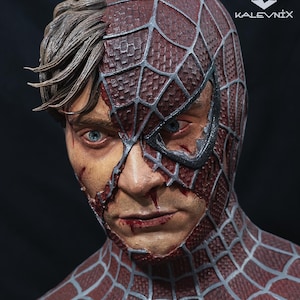 May include: A detailed bust sculpture of a human face partially covered by a red and silver spider-web patterned mask. The exposed skin shows realistic textures, blue eyes, and brown hair. The text KALEVNIX is printed in the upper right corner.