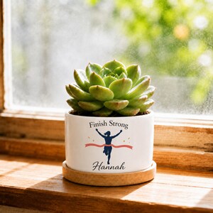 以下が含まれることがあります： 緑色の多肉植物が入った白い陶器製の植木鉢。鉢には「Finish Strong」という文字と、赤いフィニッシュテープを切るランナーのグラフィックが描かれており、その下に黒い筆記体で「Hannah」という名前がプリントされています。