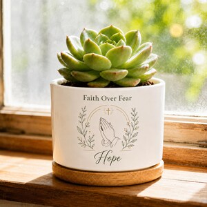 May include: A white ceramic planter containing a green succulent plant. The planter features the text Faith Over Fear and Hope, along with a graphic illustration of praying hands, a small cross, and leafy branches in a dark gray ink color.