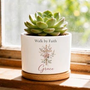 Peut inclure: Un pot de fleurs en céramique blanche avec une base en bois contenant une plante succulente verte. Le pot porte l'inscription "Walk by Faith" au-dessus d'un motif de croix orné d'illustrations florales et du mot "Grace" en écriture cursive.