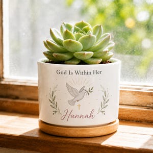 Peut inclure: Un pot de fleurs en céramique blanche contenant une plante succulente verte. Le pot porte l'inscription "God Is Within Her" au-dessus d'un motif de colombe avec un rameau d'olivier, une croix dorée et le prénom "Hannah" sur un socle en bois.