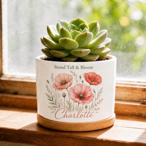 Puede incluir: Una maceta de cerámica blanca con base de madera que contiene una suculenta verde. La maceta presenta una ilustración floral de amapolas rosas, el texto "Stand Tall & Bloom" en negro y el nombre "Charlotte" en una fuente de escritura cursiva.