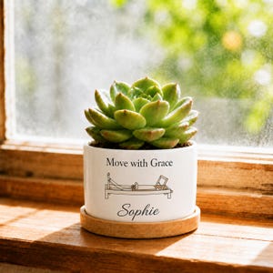 May include: A white ceramic cylindrical planter containing a green succulent plant. The planter features the black text 'Move with Grace' above a line drawing of a pilates reformer machine and the name 'Sophie' in a cursive script font below.
