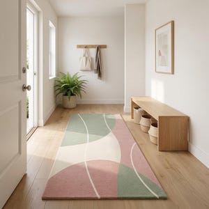 May include: A rectangular area rug featuring a geometric pattern of soft sage green and dusty rose pink curved shapes separated by thin white lines. The rug is positioned on a light-colored wooden floor in a hallway, showcasing a flat-weave texture.