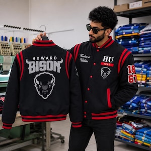 Op de afbeelding: Een zwart varsity jack met rode gestreepte boorden bij de kraag, mouwen en zoom. De achterkant toont de tekst HOWARD BISON en een afbeelding van een bizonkop. De voorkant bevat HU 1867, een bizonkop en de tekst Veritas et Utilitas in wit.